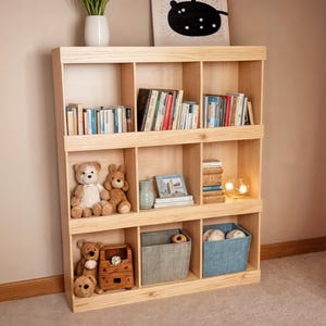 May include: A light-colored wooden bookshelf with multiple cubby shelves. The shelves are filled with books, stuffed animals, decorative items, and storage baskets. A vase with greenery and a framed picture are on display.