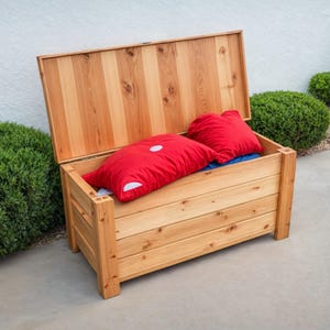 Op de afbeelding: Een houten opbergkist met een open deksel, met daarin twee rode kussens met witte accenten. De kist is gemaakt van lichtgekleurd hout en staat op een betonnen oppervlak, met groene struiken op de achtergrond.