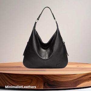 May include: A black leather hobo bag with a curved design. The bag has a single shoulder strap and a zipper closure. The bag is sitting on a wooden surface. The text "MinimalistLeathers" is at the bottom.