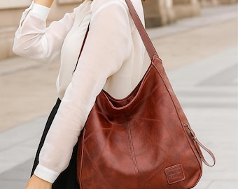May include: A brown leather shoulder bag with a long strap. The bag has a large, curved shape and a small rectangular tag on the front. The bag is worn over the shoulder. The text "MinimalistLeathers" is at the bottom.