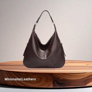 May include: A dark brown leather hobo bag with a single shoulder strap and silver-tone hardware. The bag has a curved shape and a minimalist design, with the text "MinimalistLeathers" at the bottom.