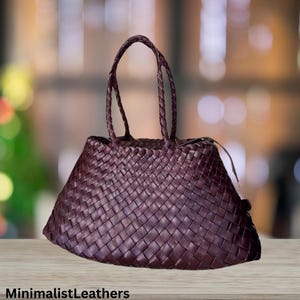 Puede incluir: Bolso tote de cuero burdeos con diseño tejido. El bolso tiene asas trenzadas y forma trapezoidal. El texto "MinimalistLeathers" está en la parte inferior de la imagen.