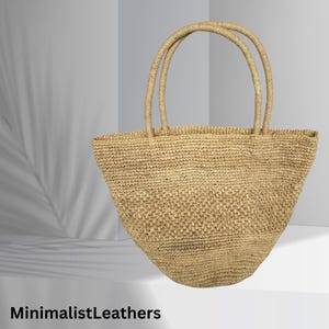 Può includere: Una borsa tote in paglia intrecciata di colore naturale con due manici arrotondati. La borsa ha una parte superiore ampia e aperta. La borsa è appoggiata su una superficie bianca. Il testo "MinimalistLeathers" è in basso nell'immagine.