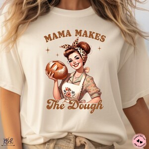 May include: A white t-shirt with a brown graphic of a woman holding a loaf of bread. The text "Mama Makes The Dough" is printed in brown letters.