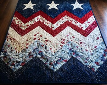 May include: A patriotic quilt with a navy blue top border featuring three white stars. The quilt showcases a red and white chevron pattern, with a floral print and a navy blue bottom border. The quilt is made of fabric and has a quilted design.