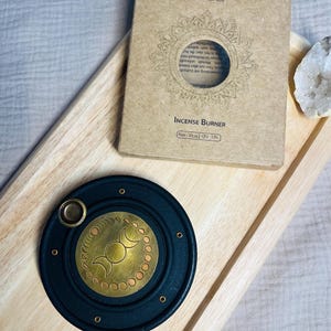 May include: A round black incense burner with a brass inlay featuring a triple moon phase design. The burner is accompanied by a rectangular cardboard box labeled Incense Burner with a 10cm size specification and a white crystal cluster.