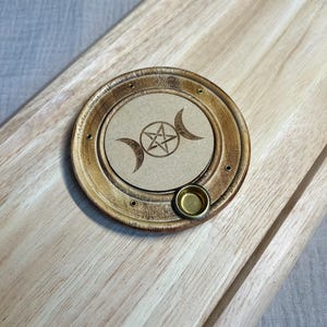 Puede incluir: Un quemador de incienso redondo de madera con un disco central beige grabado con un pentagrama flanqueado por dos lunas crecientes. El quemador tiene un acabado de madera oscura, cuatro orificios para incienso y una pequeña copa de latón.