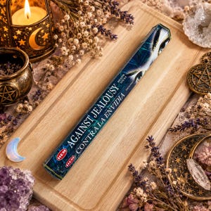 Puede incluir: Una caja rectangular de varitas de incienso HEM etiquetada como Against Jealousy y Contra la Envidia. El empaque presenta un diseño gráfico en azul oscuro y negro con una ilustración de un ojo, colocado en diagonal sobre una tabla de madera clara.
