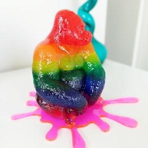 May include: A colorful gorilla sculpture in a rainbow gradient of red, orange, yellow, green, and blue. The gorilla sits on a bright pink, splattered base. A teal, twisted sculpture is in the background.