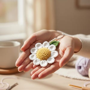 Op de afbeelding: Een gehaakte sleutelhanger in de vorm van een madeliefje met een gele kern en witte bloemblaadjes. De sleutelhanger heeft een groen blaadje en een zilverkleurige ring.