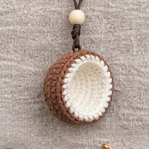 May include: A handmade crochet coconut pendant. The pendant features a brown exterior with a white interior, and a brown cord with a wooden bead. The coconut is suspended against a neutral background.