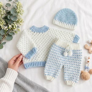 Può includere: Un completo per neonati all'uncinetto bianco e azzurro, composto da un maglione, pantaloni e un berretto. Il maglione e i pantaloni presentano dettagli blu. Il berretto è azzurro e bianco. Il set è presentato su una superficie bianca.