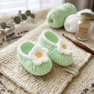 Può includere: Un paio di scarpine per neonati lavorate all'uncinetto in verde chiaro, con un'applicazione a forma di fiore bianco e giallo sulla parte anteriore di ogni scarpa. Le scarpine presentano un punto testurizzato e un bordo bianco.