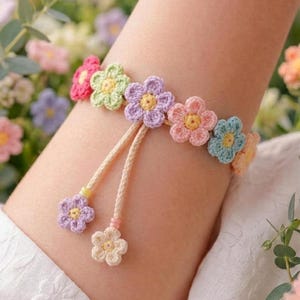 Op de afbeelding: Een kleurrijke gehaakte bloemenarmband. De armband heeft gehaakte bloemen in roze, groen, paars en blauw. De armband is bevestigd aan een beige koord met twee bungelende bloembedels.