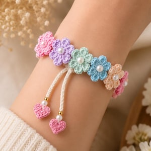 May include: A crochet bracelet featuring a row of multi-colored flowers in pink, lavender, mint green, blue, and peach, each centered with a small white pearl. Two cream-colored cords hang down, each finished with a small pink crochet heart and a pearl.