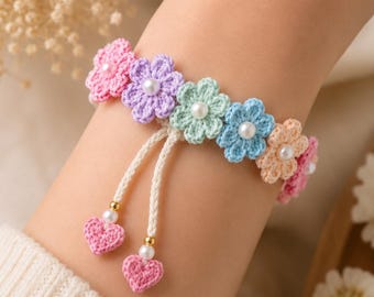 Può includere: Un braccialetto all'uncinetto con una fila di fiori multicolori in rosa, lavanda, verde menta, blu e pesca, ognuno con una piccola perla bianca al centro. Due cordini color crema pendono verso il basso, terminati da un piccolo cuore all'uncinetto rosa.