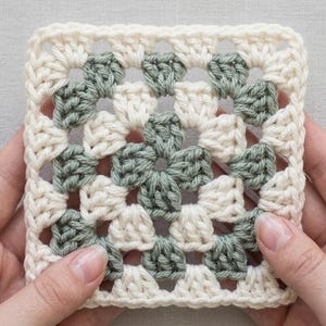 May include: A square, handmade crochet granny square in cream and sage green. The square is held by two hands. The crochet pattern features a floral design in the center and corners, with a solid cream border.