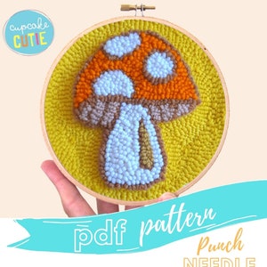 May include: A punch needle pattern for a mushroom with a brown stem, orange and white cap, and white dots. The pattern is in a wooden embroidery hoop on a yellow background. The text "pdf pattern Punch NEEDLE" is on a blue ribbon.