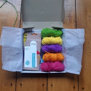 May include: A box filled with colorful yarn for crafting. The yarn is in shades of green, yellow, purple, orange, and pink. The box also includes a small card with the word "LOOME" printed on it.