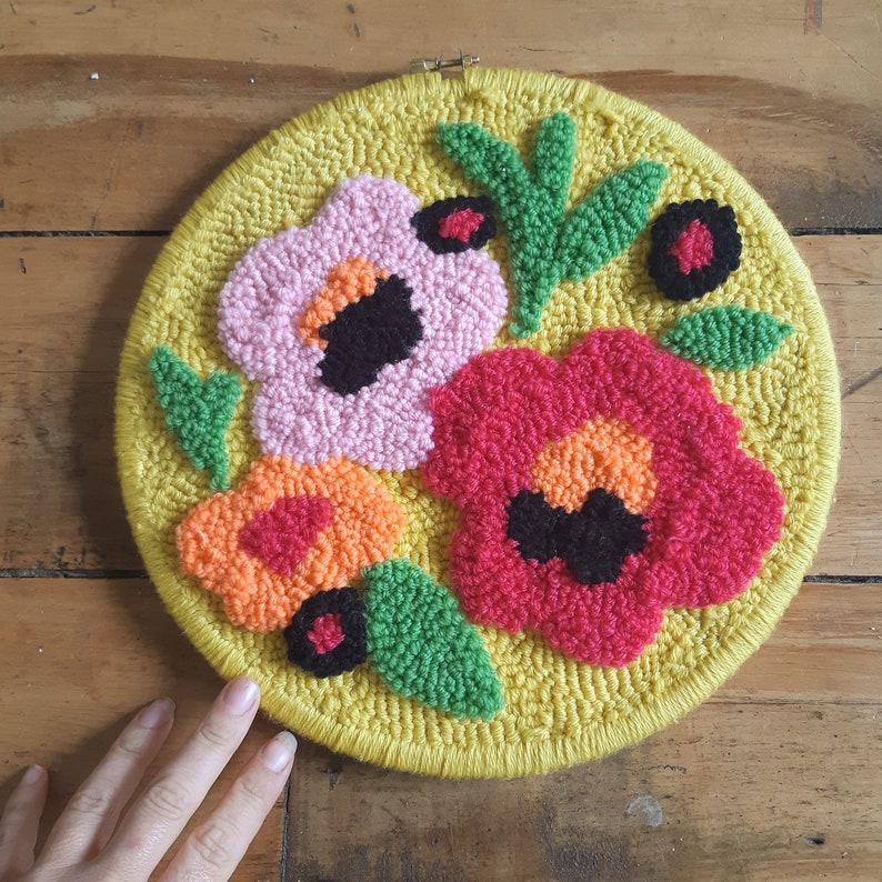 Op de afbeelding: Een ronde, gele borduurring met een bloemenmotief. Het ontwerp bevat drie bloemen in roze, oranje en rood met zwarte centra. De bloemen worden omringd door groene bladeren.