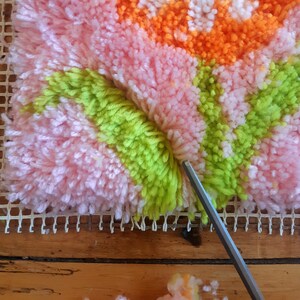 May include: Close-up of a pink, green, and orange rug being made with a rug hooking tool. The rug is being made on a white canvas grid.