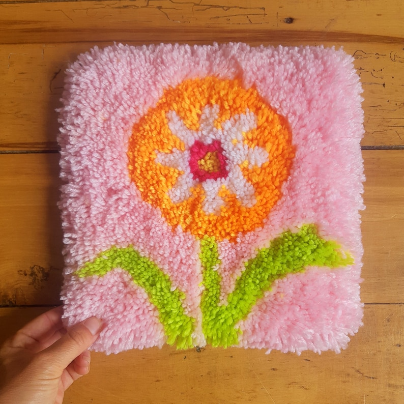 May include: A pink, orange, white, and green rug with a flower design. The flower has a white center with a red and orange center. The flower is surrounded by green leaves.