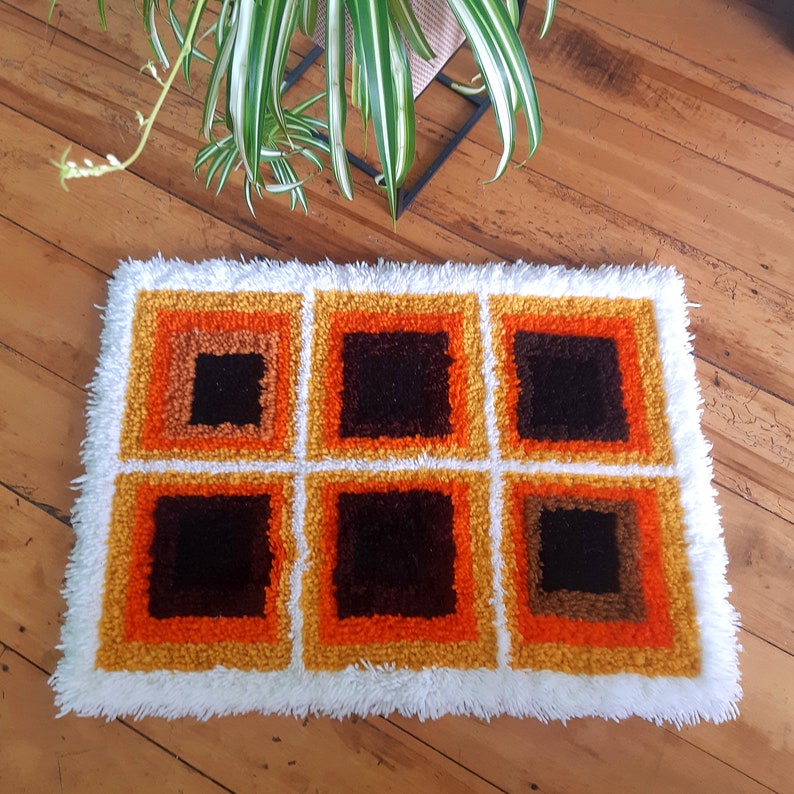 May include: A white shag rug with a geometric pattern of orange, brown, and black squares. The rug has a white fringe border.