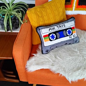 May include: An orange armchair with a white faux fur throw and two pillows. One pillow is a yellow knitted pillow and the other is a gray pillow with a needlepoint design of a cassette tape with the text "Mix Tape" and "FE 90".