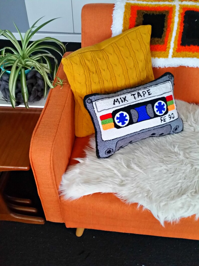 May include: An orange armchair with a white faux fur throw and two pillows. One pillow is a yellow knitted pillow and the other is a gray pillow with a needlepoint design of a cassette tape with the text "Mix Tape" and "FE 90".