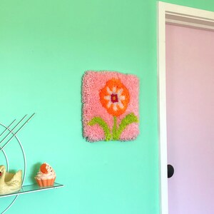 May include: A pink and orange flower rug with green leaves hangs on a light green wall. The rug is made of yarn and has a fluffy texture.