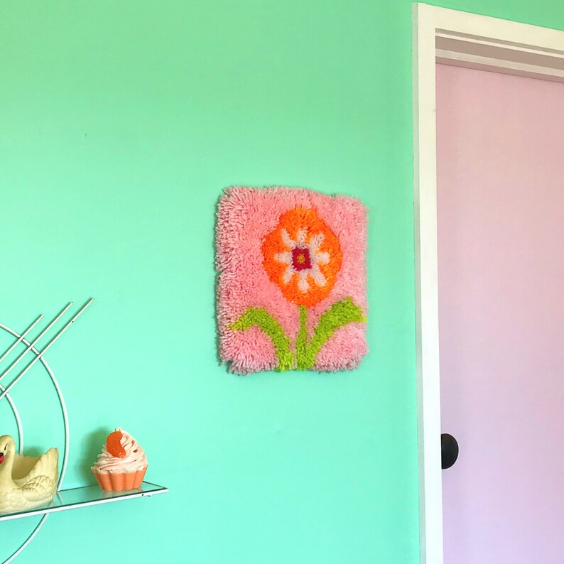 May include: A pink and orange flower rug with green leaves hangs on a light green wall. The rug is made of yarn and has a fluffy texture.