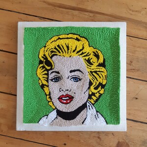 Puede incluir: Una obra de arte de punto de aguja enmarcada que representa a una mujer con cabello rubio y una camisa blanca sobre un fondo verde.