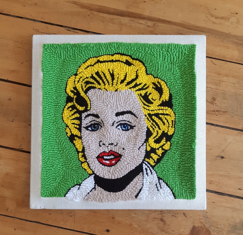 Puede incluir: Una obra de arte de punto de aguja enmarcada que representa a una mujer con cabello rubio y una camisa blanca sobre un fondo verde.