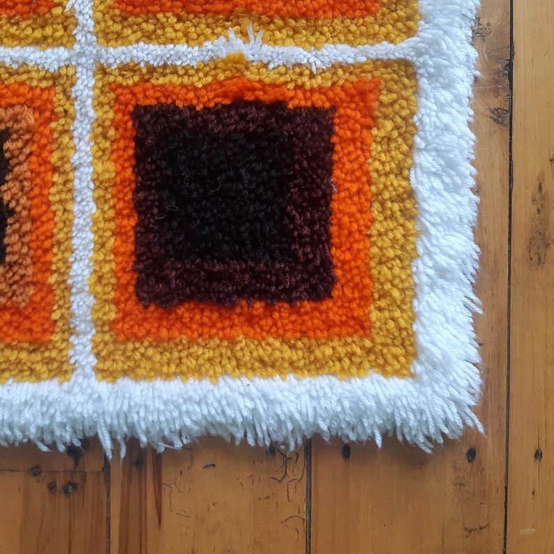 May include: A close-up of a colorful, geometric rug with a white fringe. The rug features squares of orange, yellow, and brown.