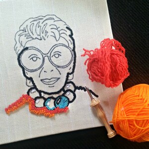 Op de afbeelding: Een borduurportret van een vrouw met een grote bril, in uitvoering. Het ontwerp is in zwart omlijnd op een beige canvas. Een klein deel van het portret is gevuld met oranje, blauw en wit garen. Twee bollen garen, &eacute;&eacute;n oranje en &eacute;&eacute;n rood, en een punch needle zijn ook zichtbaar.