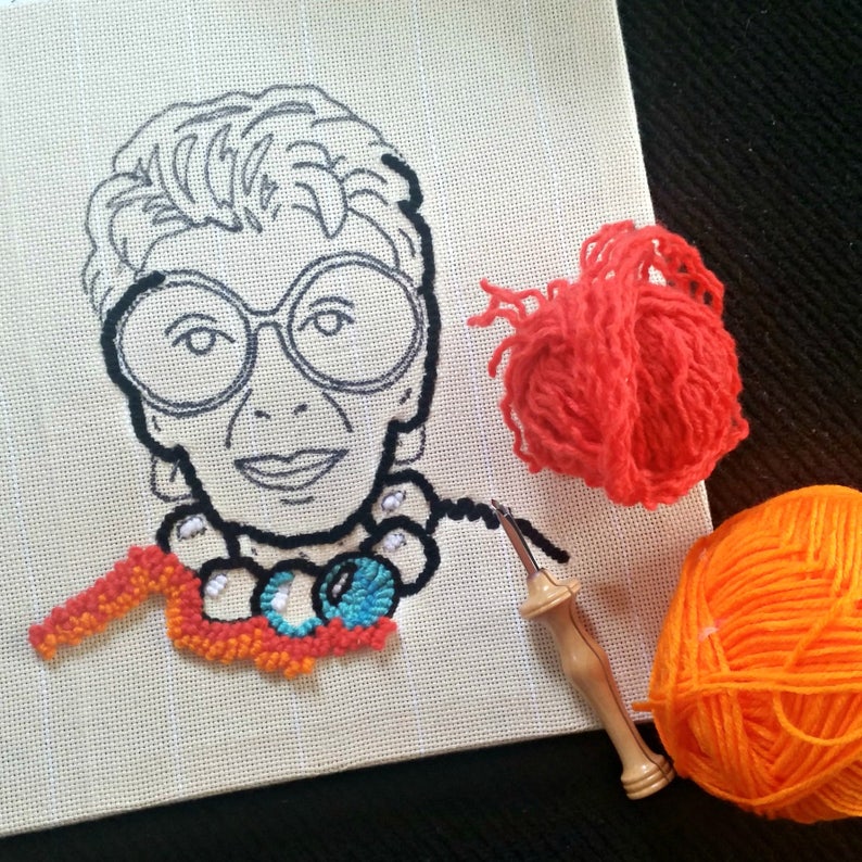 Op de afbeelding: Een borduurportret van een vrouw met een grote bril, in uitvoering. Het ontwerp is in zwart omlijnd op een beige canvas. Een klein deel van het portret is gevuld met oranje, blauw en wit garen. Twee bollen garen, &eacute;&eacute;n oranje en &eacute;&eacute;n rood, en een punch needle zijn ook zichtbaar.