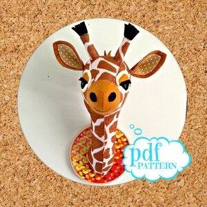 Puede incluir: Una cabeza de jirafa de fieltro para colgar en la pared con un diseño estampado en marrón y blanco. La jirafa tiene una expresión amigable y está montada en un círculo con un patrón geométrico colorido. El texto "pdf pattern" está escrito en una burbuja de pensamiento azul.