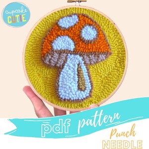 May include: A punch needle pattern for a mushroom with a brown stem, orange and white cap, and white spots. The pattern is on a yellow background and is in a wooden embroidery hoop.