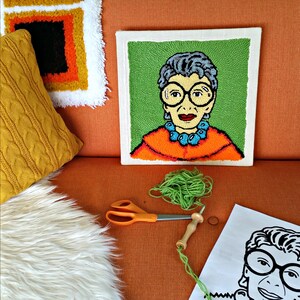 Op de afbeelding: Een kleurrijk, handgemaakt punch needle kunstwerk met een portret van een vrouw met een bril en een turquoise ketting. Het kunstwerk staat op een vierkant canvas. Groen garen, een schaar en een punch needle tool zijn zichtbaar.