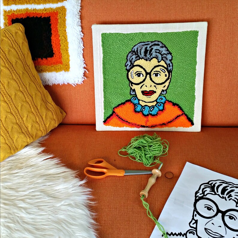 Op de afbeelding: Een kleurrijk, handgemaakt punch needle kunstwerk met een portret van een vrouw met een bril en een turquoise ketting. Het kunstwerk staat op een vierkant canvas. Groen garen, een schaar en een punch needle tool zijn zichtbaar.