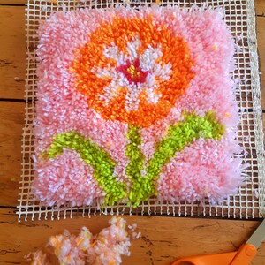 May include: A pink, orange, white, and green rug hooking project of a flower with leaves. The project is on a white canvas grid and there are loose strands of yarn nearby.