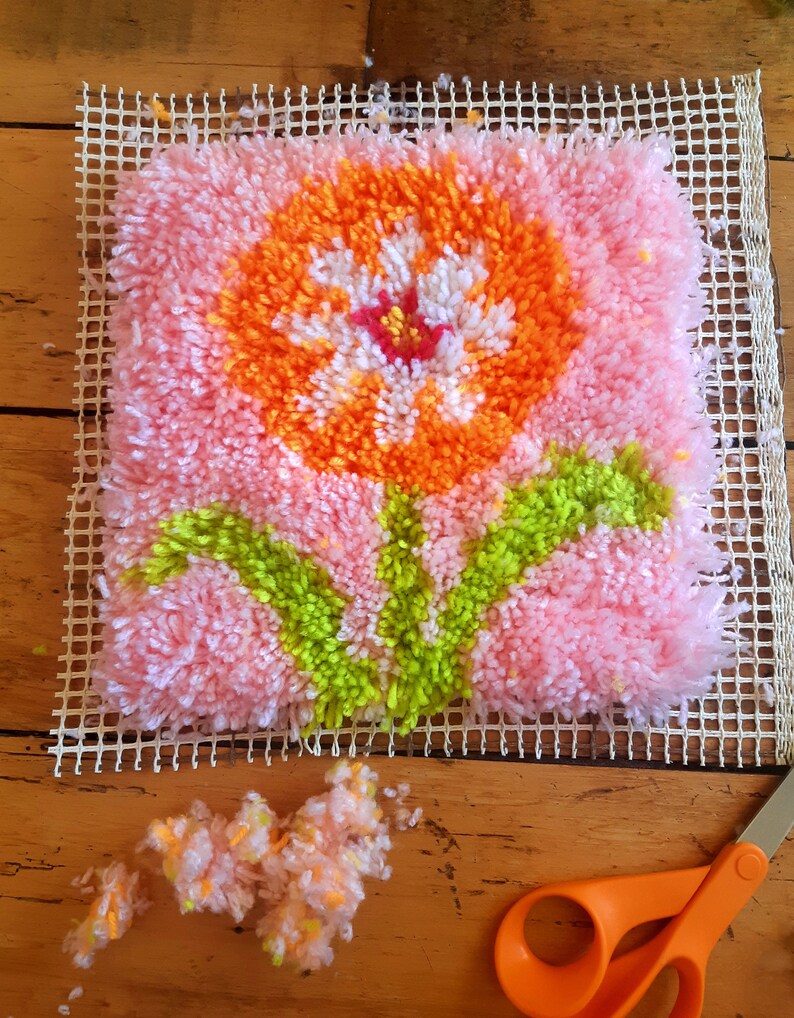 May include: A pink, orange, white, and green rug hooking project of a flower with leaves. The project is on a white canvas grid and there are loose strands of yarn nearby.