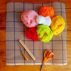 May include: A white canvas with a grid pattern, used for needlepoint, is covered with colorful yarn balls in shades of pink, orange, yellow, green, and red. A wooden needle tool and a pair of silver scissors are also on the canvas.