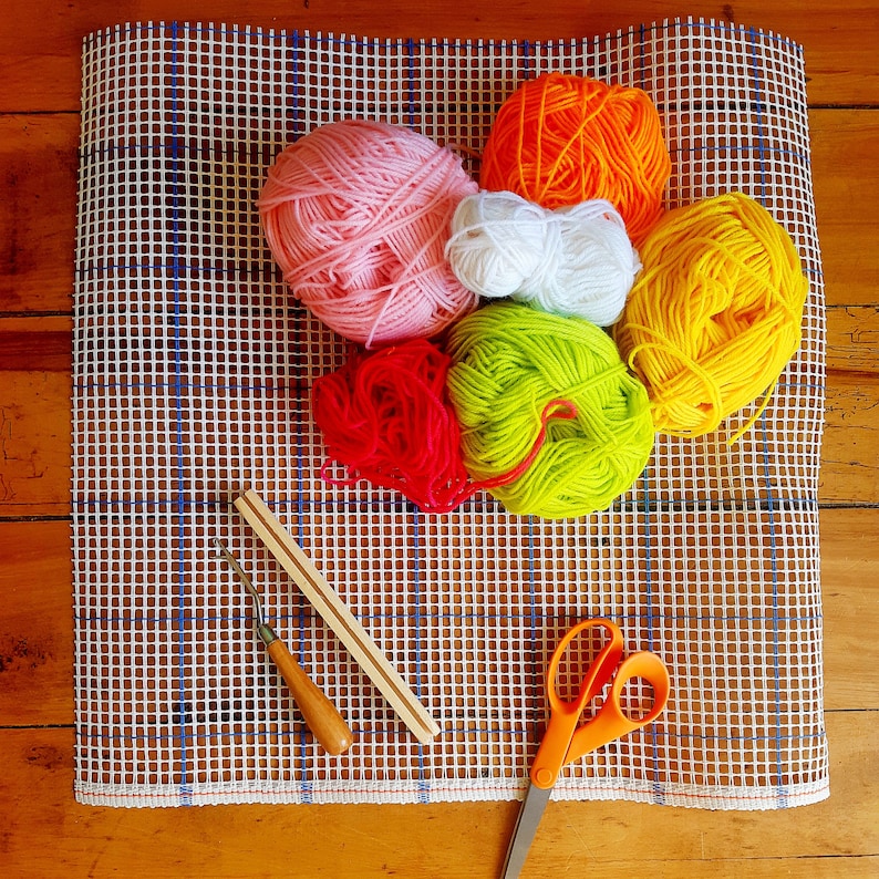 May include: A white canvas with a grid pattern, used for needlepoint, is covered with colorful yarn balls in shades of pink, orange, yellow, green, and red. A wooden needle tool and a pair of silver scissors are also on the canvas.
