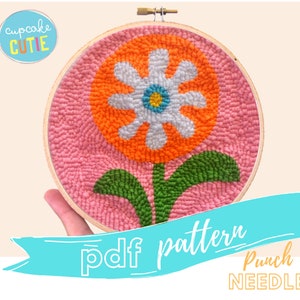 May include: A punch needle embroidery pattern featuring a large orange flower with a white center and green leaves on a pink background. The pattern is in a hoop and the text "pdf pattern Punch NEEDLE" is below the hoop.