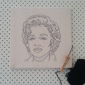 Puede incluir: Un dibujo en blanco y negro de la cara de una mujer sobre un lienzo blanco. El lienzo est&aacute; sobre una superficie blanca con puntos negros. Tambi&eacute;n hay una herramienta de aguja de madera y una bola de hilo negro en la imagen.