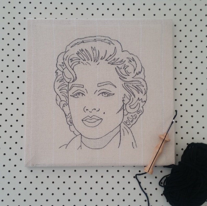 Puede incluir: Un dibujo en blanco y negro de la cara de una mujer sobre un lienzo blanco. El lienzo est&aacute; sobre una superficie blanca con puntos negros. Tambi&eacute;n hay una herramienta de aguja de madera y una bola de hilo negro en la imagen.