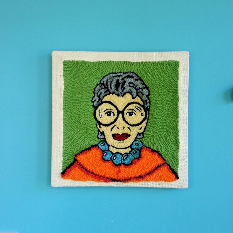 Op de afbeelding: Een vierkant, met de hand getuft wandkleed met een portret van een vrouw met een grote, ronde bril. Het kunstwerk heeft een groene achtergrond, een witte rand en de vrouw draagt een oranje top en een blauwe kralenketting. Het is een kleurrijk, getextureerd stuk.
