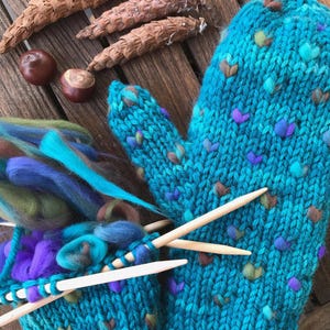 May include: A pair of turquoise knitted mittens with colorful accents, alongside knitting needles and a ball of yarn. The mittens have a textured pattern with small, heart-shaped embellishments. Natural elements like pine cones and chestnuts are arranged nearby.
