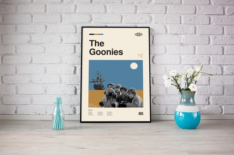 May include: A black framed poster with a minimalist design featuring a blue and yellow colour scheme. The poster has the text "The Goonies" in bold black letters and a silhouette of a ship with a group of children in front of it. The poster also includes the year "1985" in the bottom right corner.